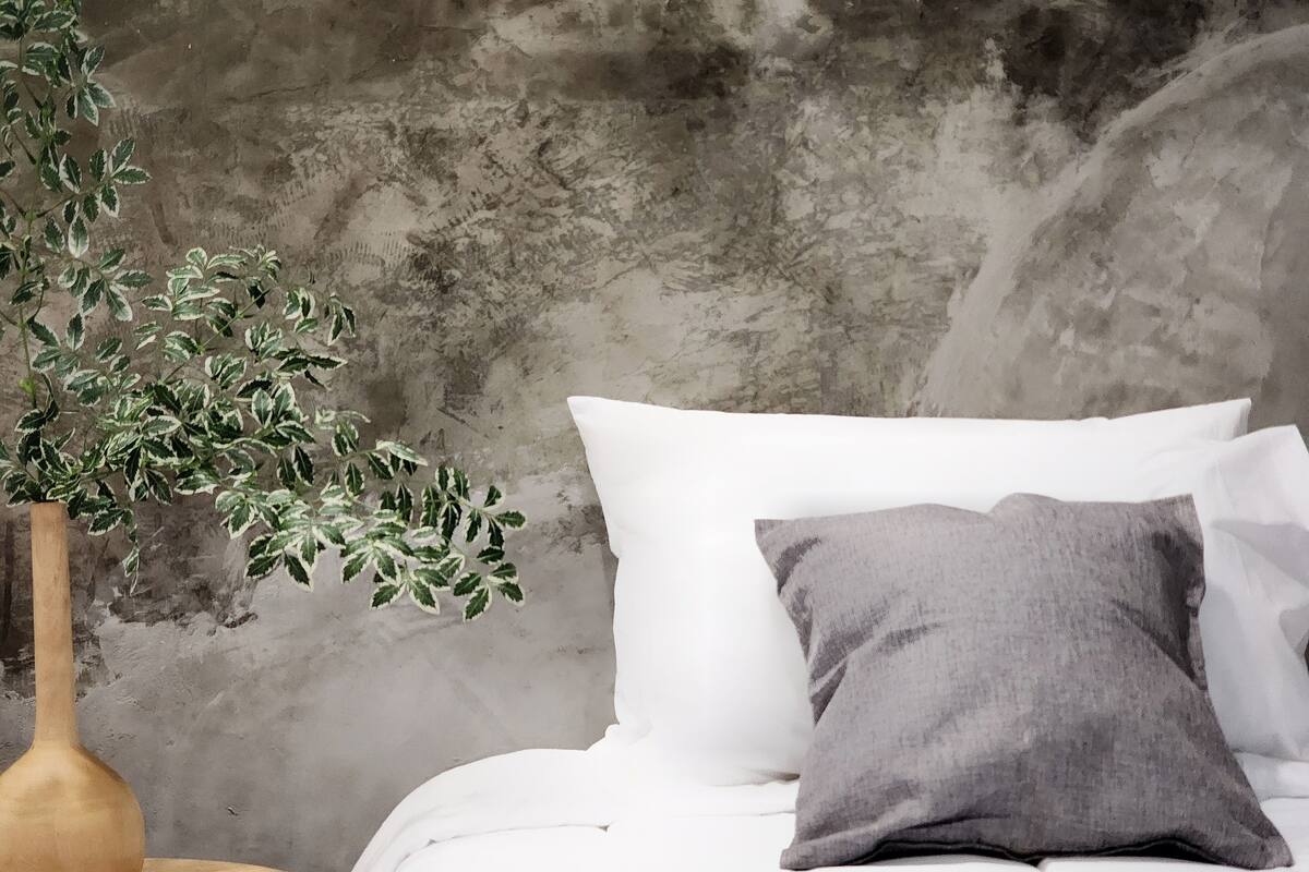 Un lit avec une literie blanche et un oreiller gris, adossé à un mur en béton à la texture particulière.