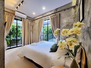 A bedroom with a large bed, beige curtains, a potted plant, and a window with a view of greenery. - Art Gate Hotel (Chiang Mai)