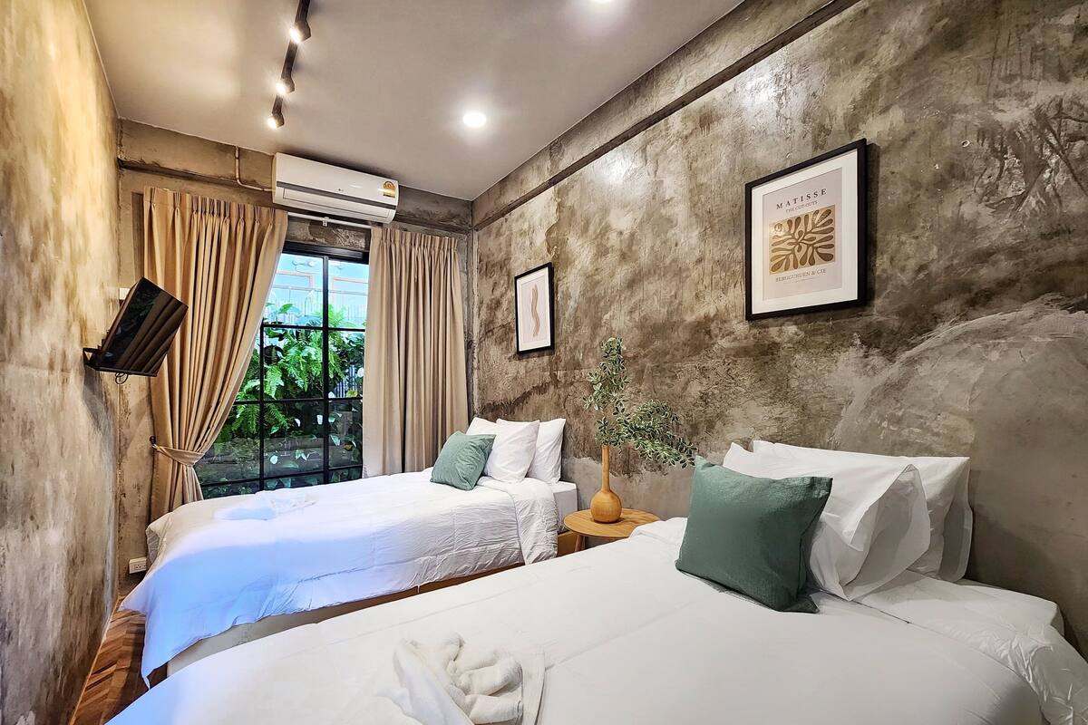 Une chambre d’hôtel avec deux lits, une grande fenêtre avec des rideaux, un climatiseur fixé au mur, un tableau encadré et une petite table avec un vase.
