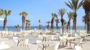 Un espacio al aire libre con mesas y sillas blancas, palmeras y vistas al mar.
