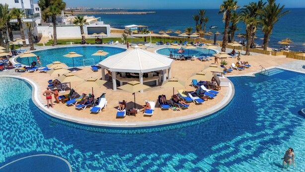 Zona de piscina en un resort con varios lugares para tomar el sol, un pabellón central y vista al mar.