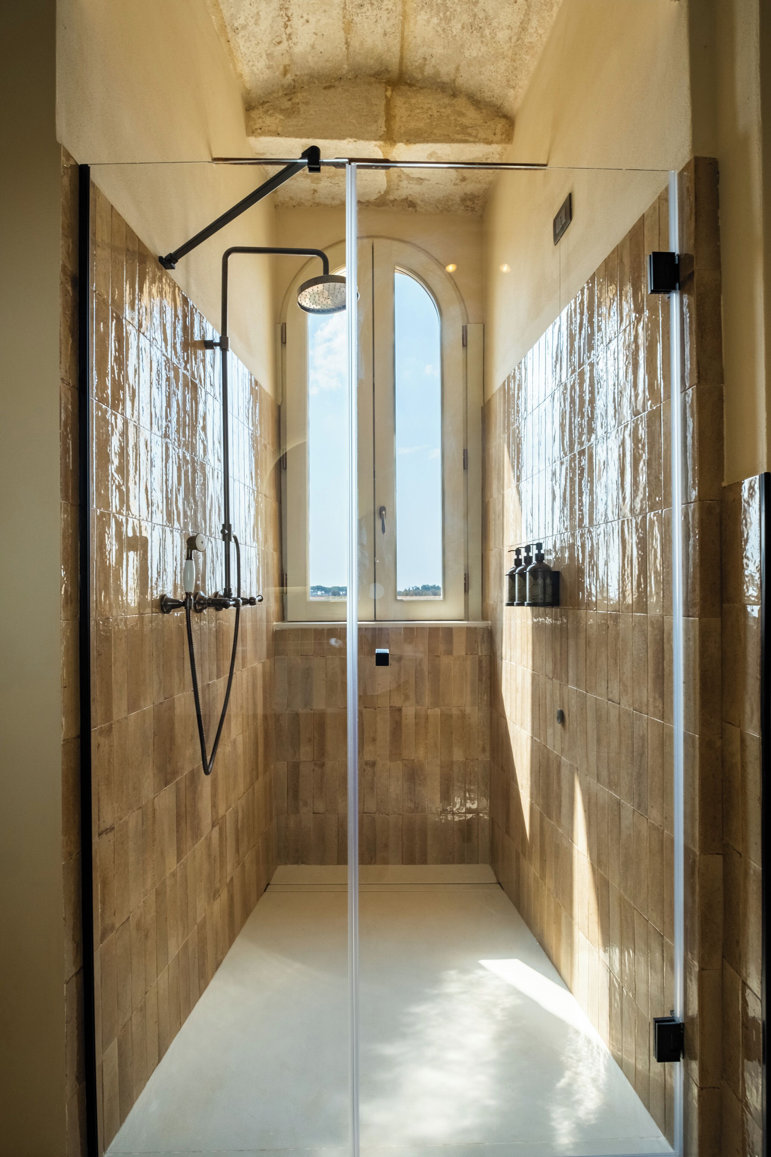 A modern bathroom with a glass shower enclosure, tiled walls, and a view through the arched window.