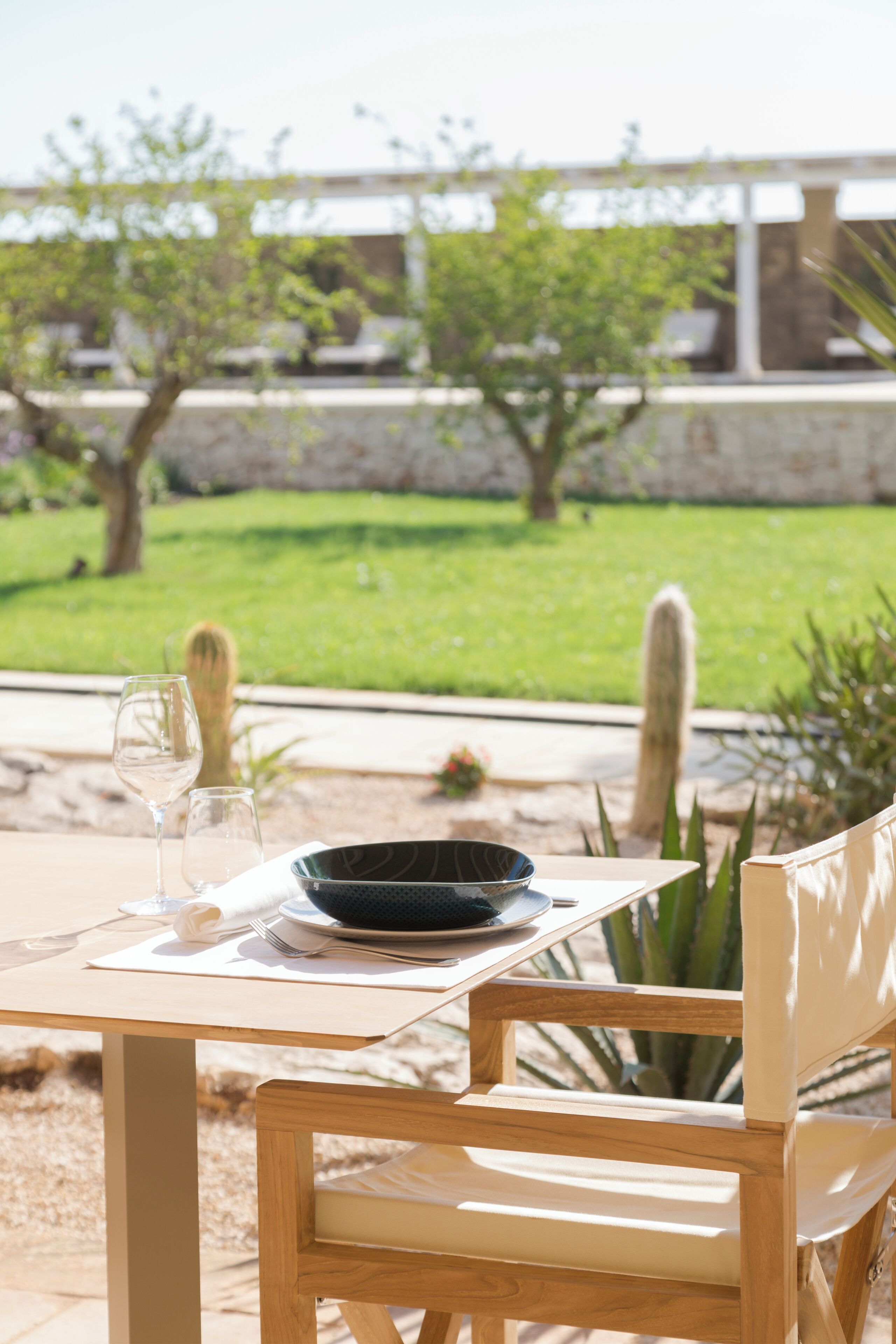 Une table dressée pour deux personnes, avec un saladier et des verres à vin, donnant sur un jardin agrémenté de cactus et d’un mur de pierre.