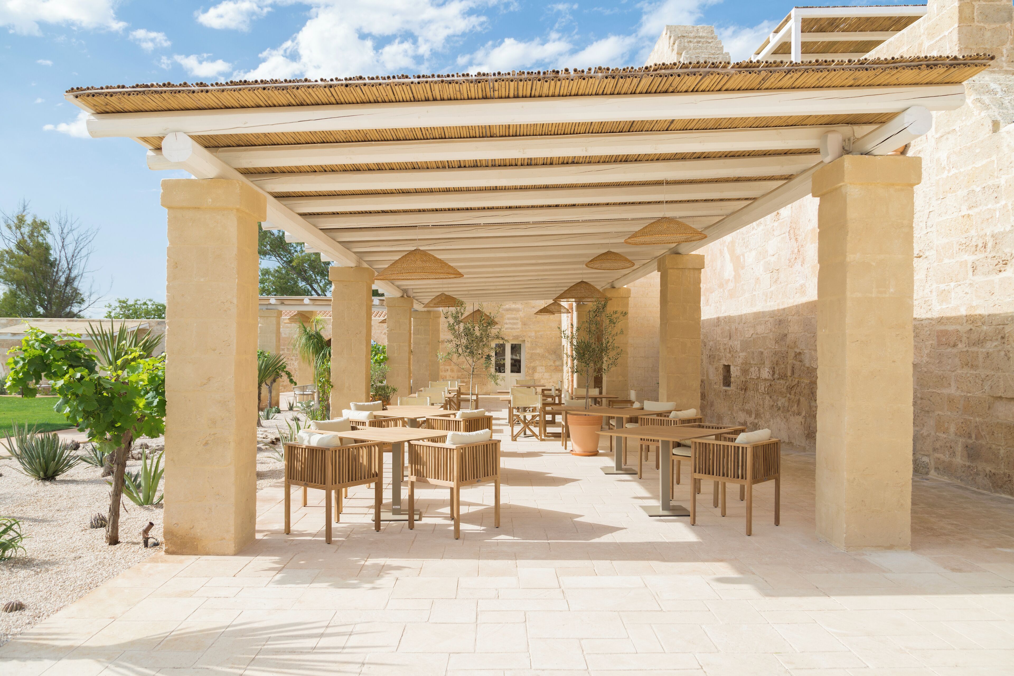 Eine Außenpatio-Terrasse mit mehreren Holztischen und Stühlen, einer Pergola und einer Mauer aus Stein im Hintergrund.