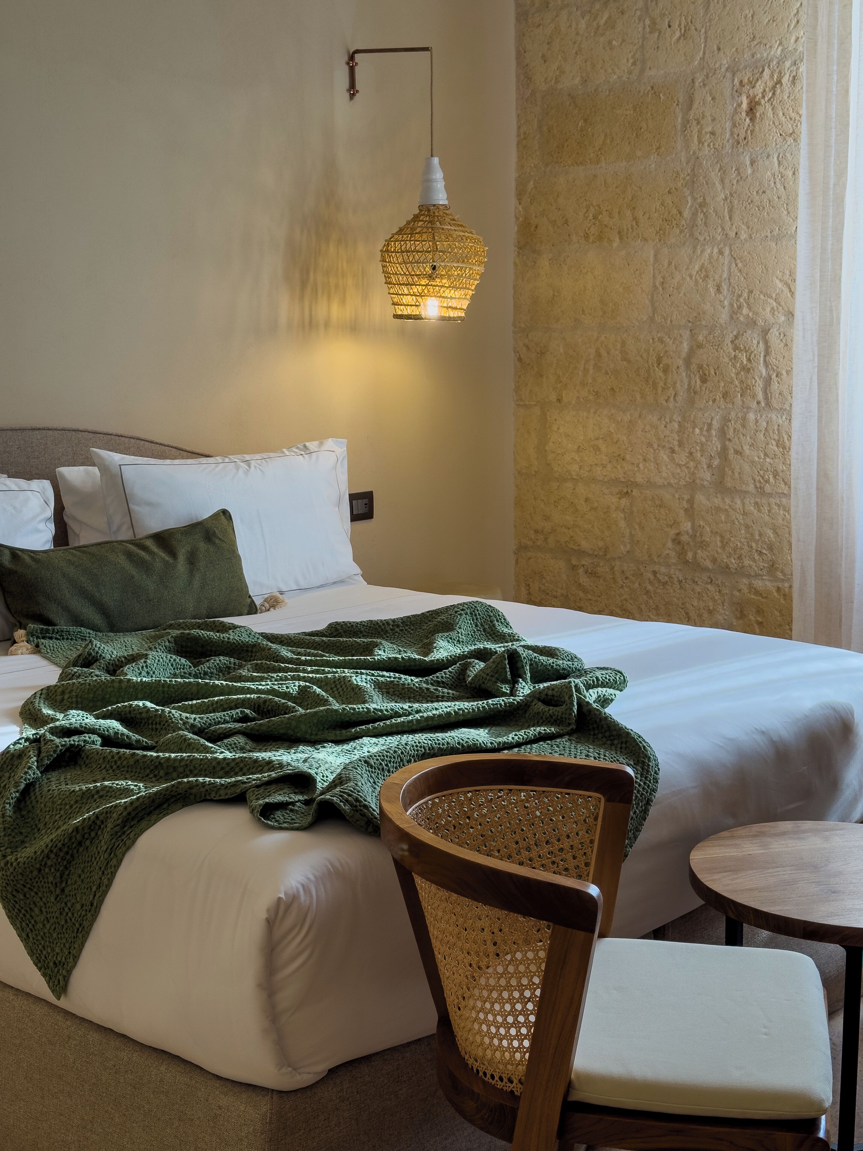 A neatly made bed with white linens and a green knitted blanket, a wooden chair with a woven seat, and a small wooden table beside the bed.