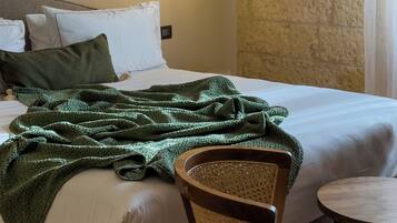A neatly made bed with white linens and a green knitted blanket, a wooden chair with a woven seat, and a small wooden table beside the bed.