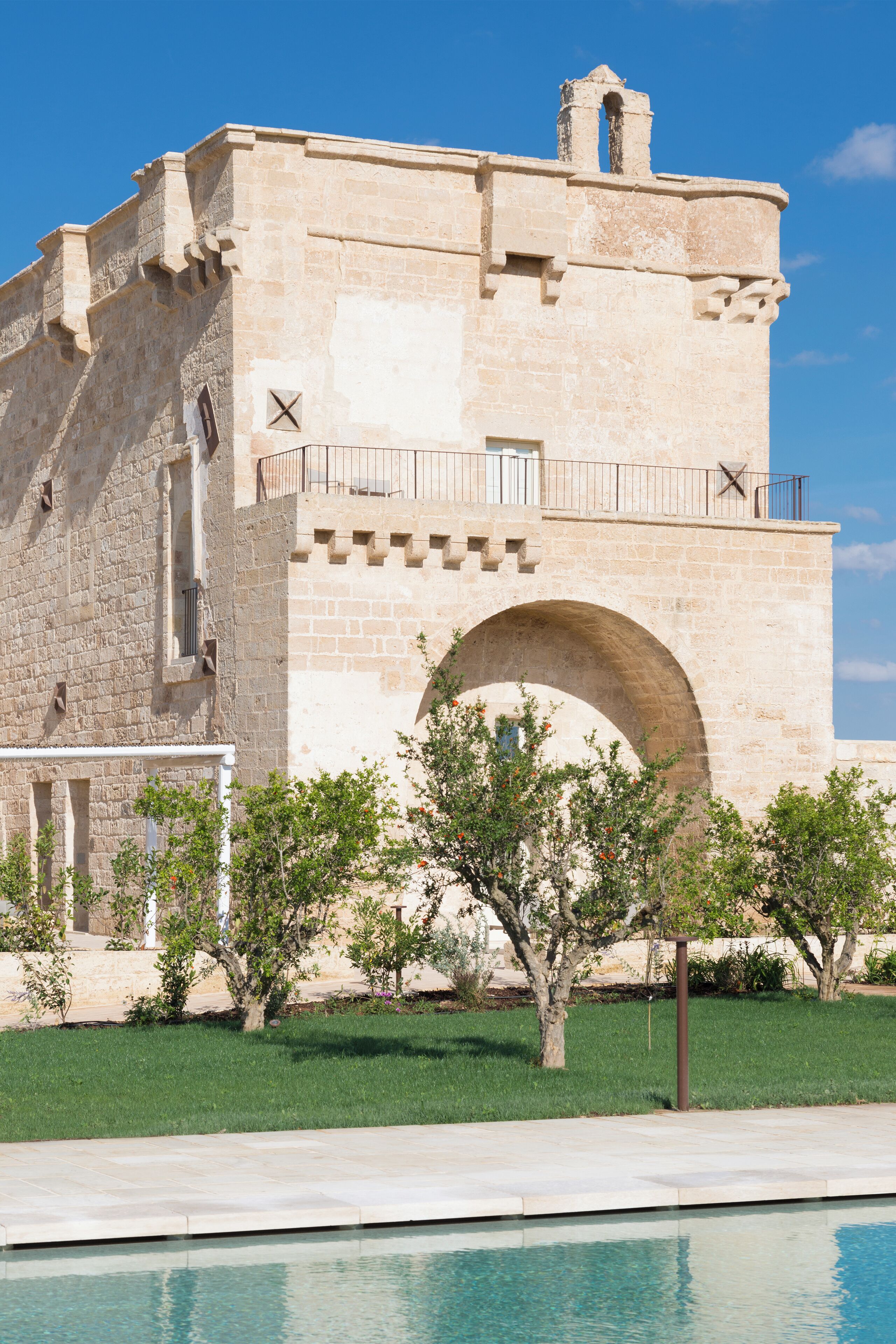 Un bâtiment historique en pierre, doté d’une grande porte en arc de cercle, d’un balcon avec une balustrade et d’une piscine en premier plan.