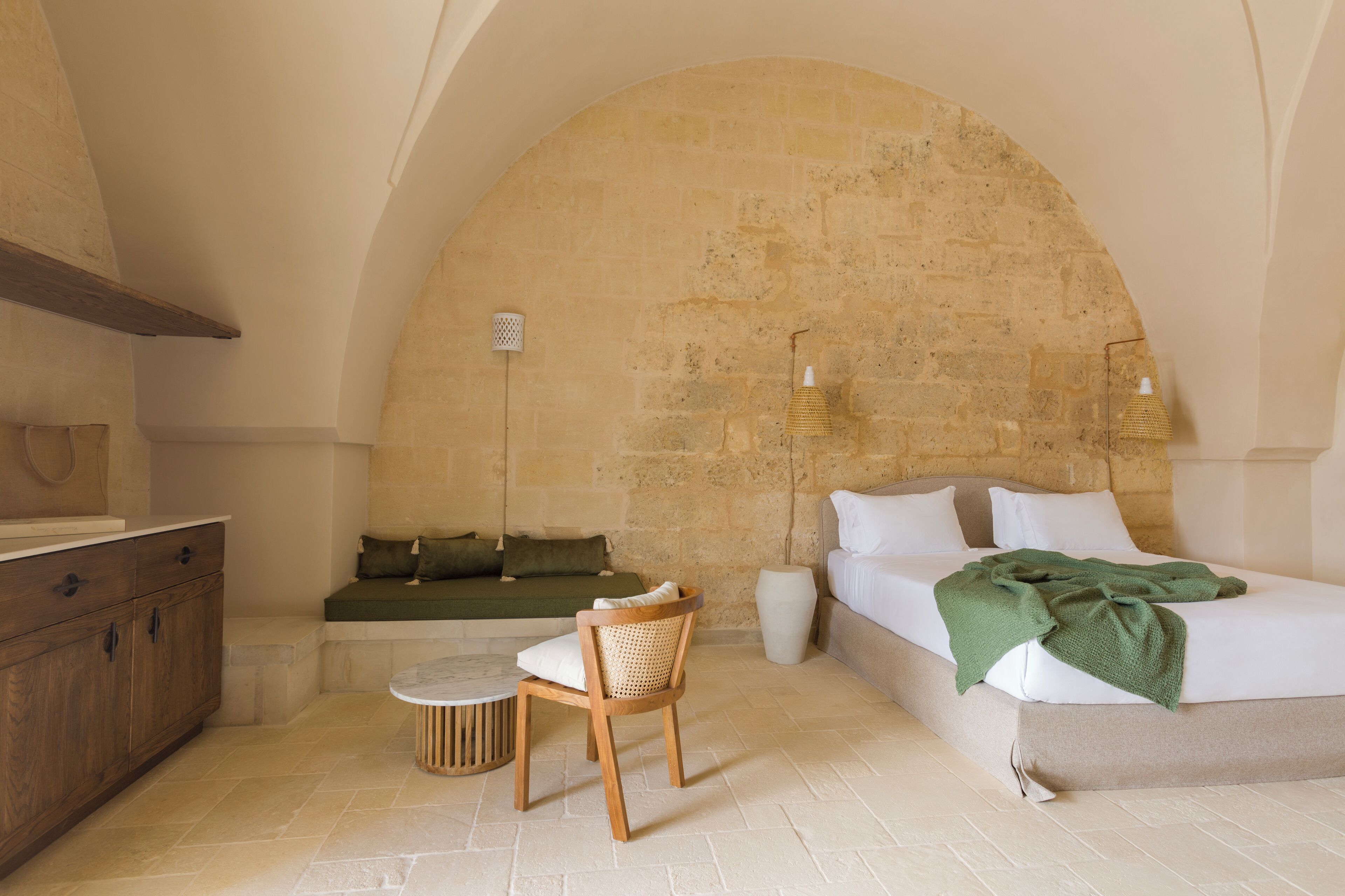 Un dormitorio con pared de piedra, una cama con manta verde, una silla de madera, una mesita redonda pequeña y una mesita de noche.