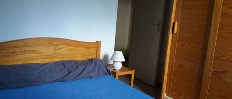 A bedroom with a wooden headboard, a blue bedspread, a white lamp on a bedside table, and a wooden wardrobe.