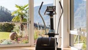 A stationary exercise bike with a digital display, positioned near a large window with a view of a garden and mountains. - Hotel Kiendl (Schenna)