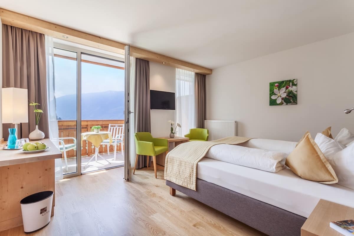 A hotel room with a bed, a desk with a lamp, a chair, a small table with a plant, and a view of the outdoors.