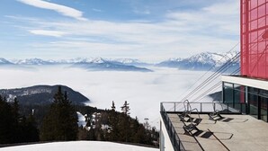 A snow-covered mountain landscape with a clear sky. A building with a red facade and glass windows is visible, featuring outdoor seating with chairs. - Hotel Kiendl (Schenna)