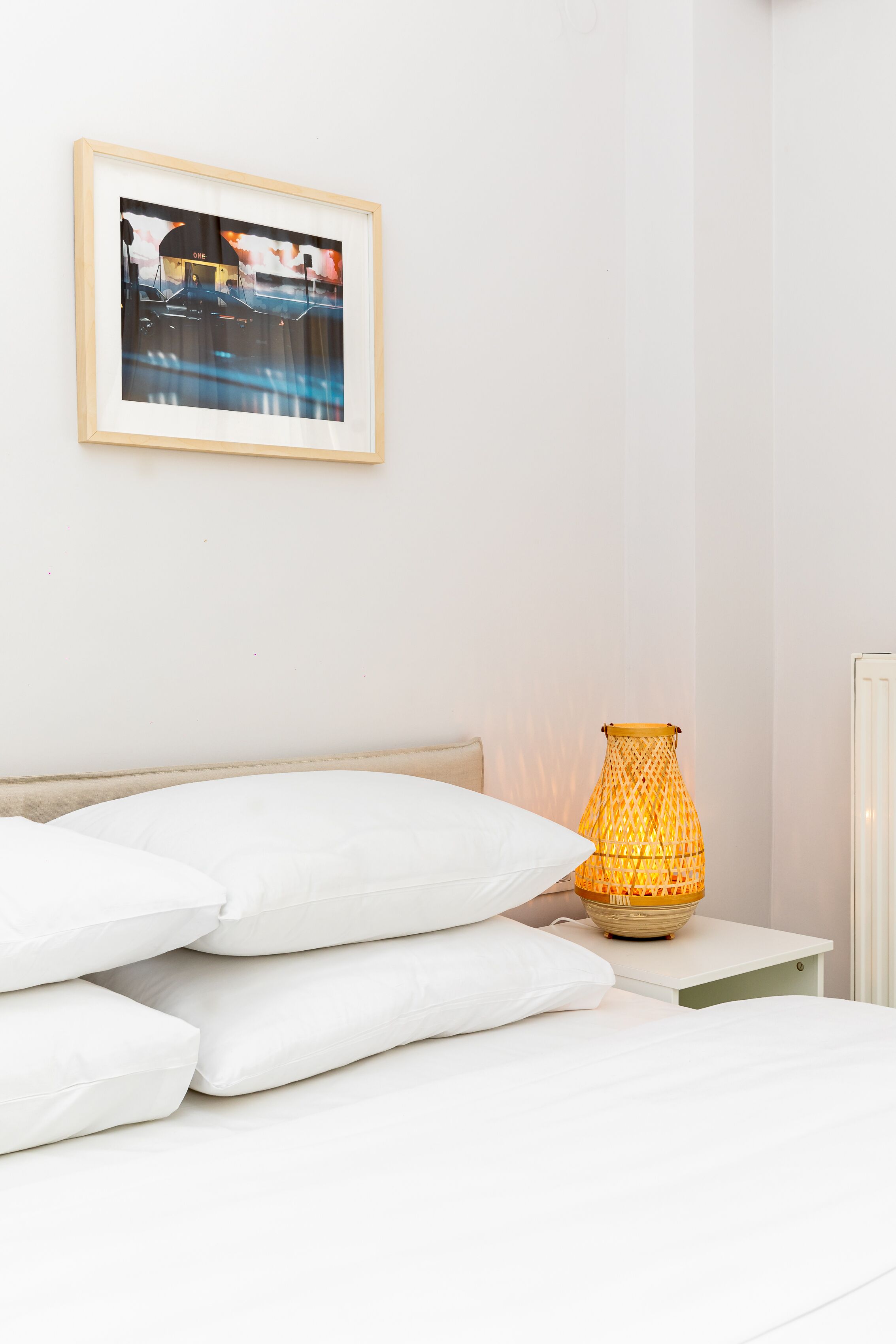 A neatly made bed with white bedding, a framed picture on the wall, and a decorative lamp on a bedside table.