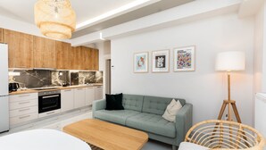 A modern kitchen with wooden cabinets, a built-in oven, and a marble backsplash. There is a light green sofa with a black pillow, a wooden table, and a wicker chair. Three framed artworks are displayed on the wall. - Sunflower by halu! Apartments (Thessaloniki)