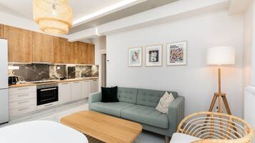 A modern kitchen with wooden cabinets, a built-in oven, and a marble backsplash. There is a light green sofa with a black pillow, a wooden table, and a wicker chair. Three framed artworks are displayed on the wall.