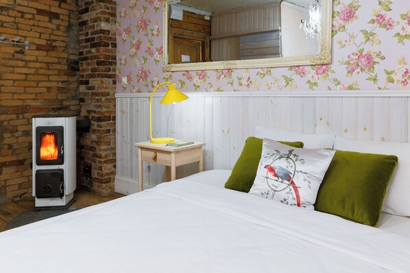 A room with a brick fireplace, a wooden nightstand, a bed with green and white pillows, and a floral wallpaper. - Marta 8 - House & Garden (Tallinn)