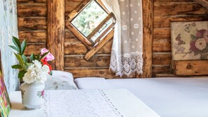 Una habitación rústica de cabaña de madera con una cama, una ventana con cortinas de encaje y un florero con flores sobre una mesa.