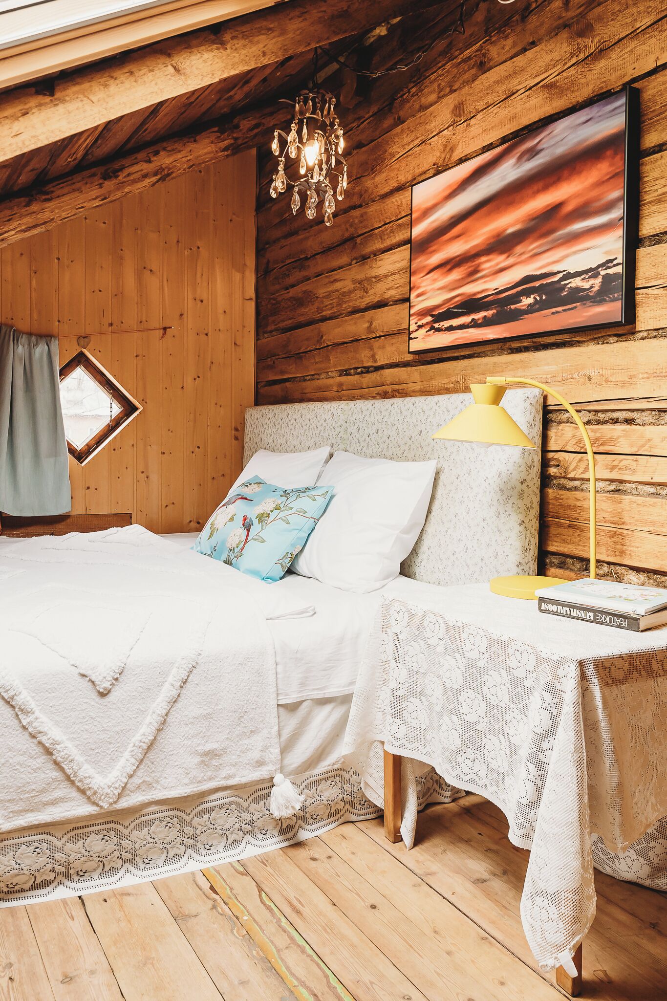 A cozy bedroom with a wooden ceiling, a bed with white linens, a small table with a yellow lamp, and a framed picture on the wall.