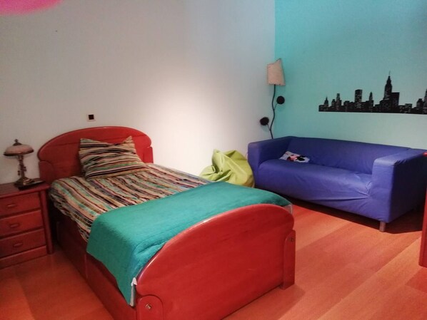 A bedroom with a red bed, a blue sofa, a cityscape wall decoration, and a lamp on a bedside table.
