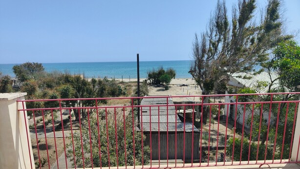 Varanda com guarda-corpo de metal vermelho, vista para uma praia e um pequeno edifício.