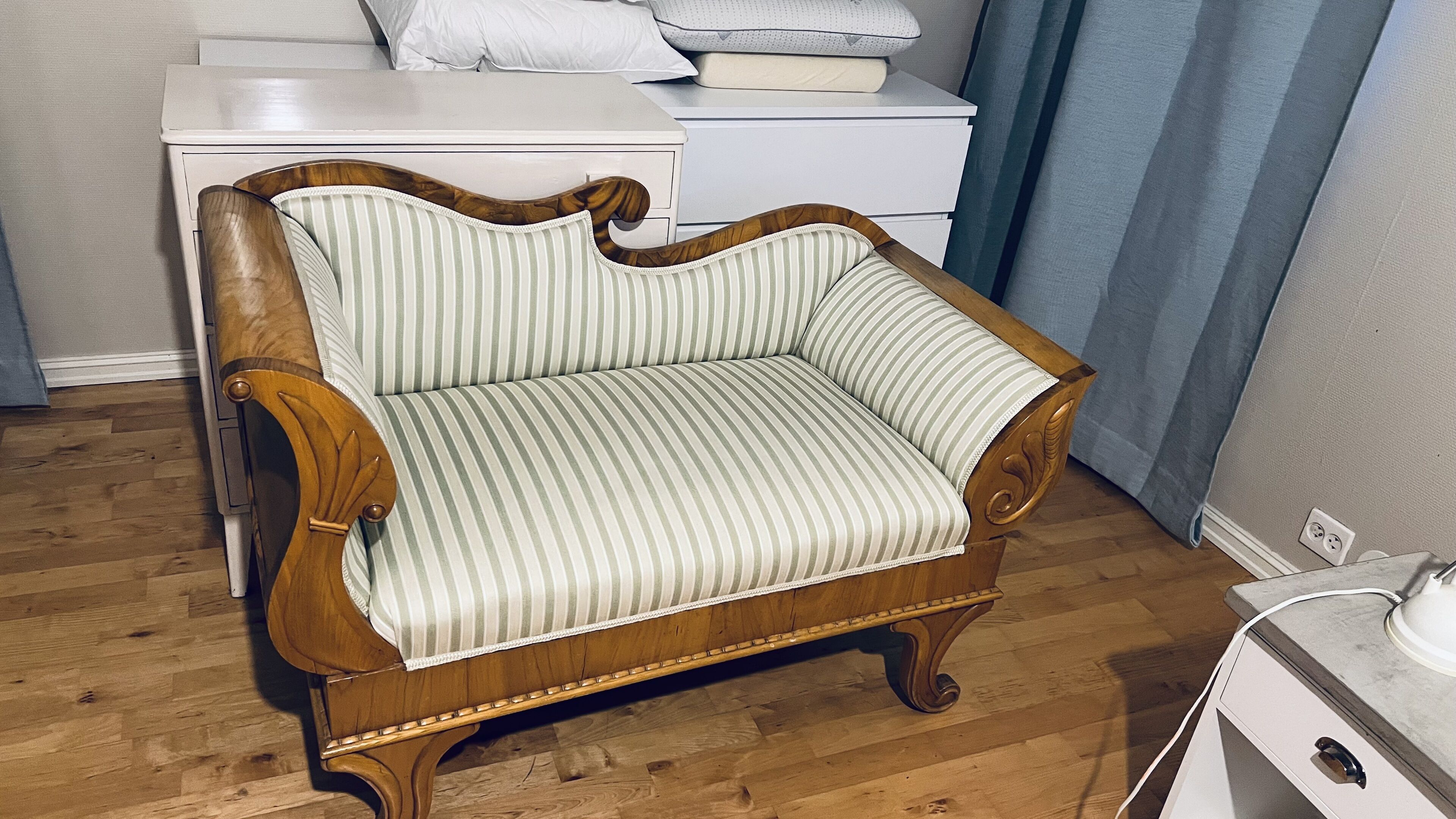 A wooden armchair with a striped cushion in a room with a wooden floor, a white dresser, and a blue curtain.