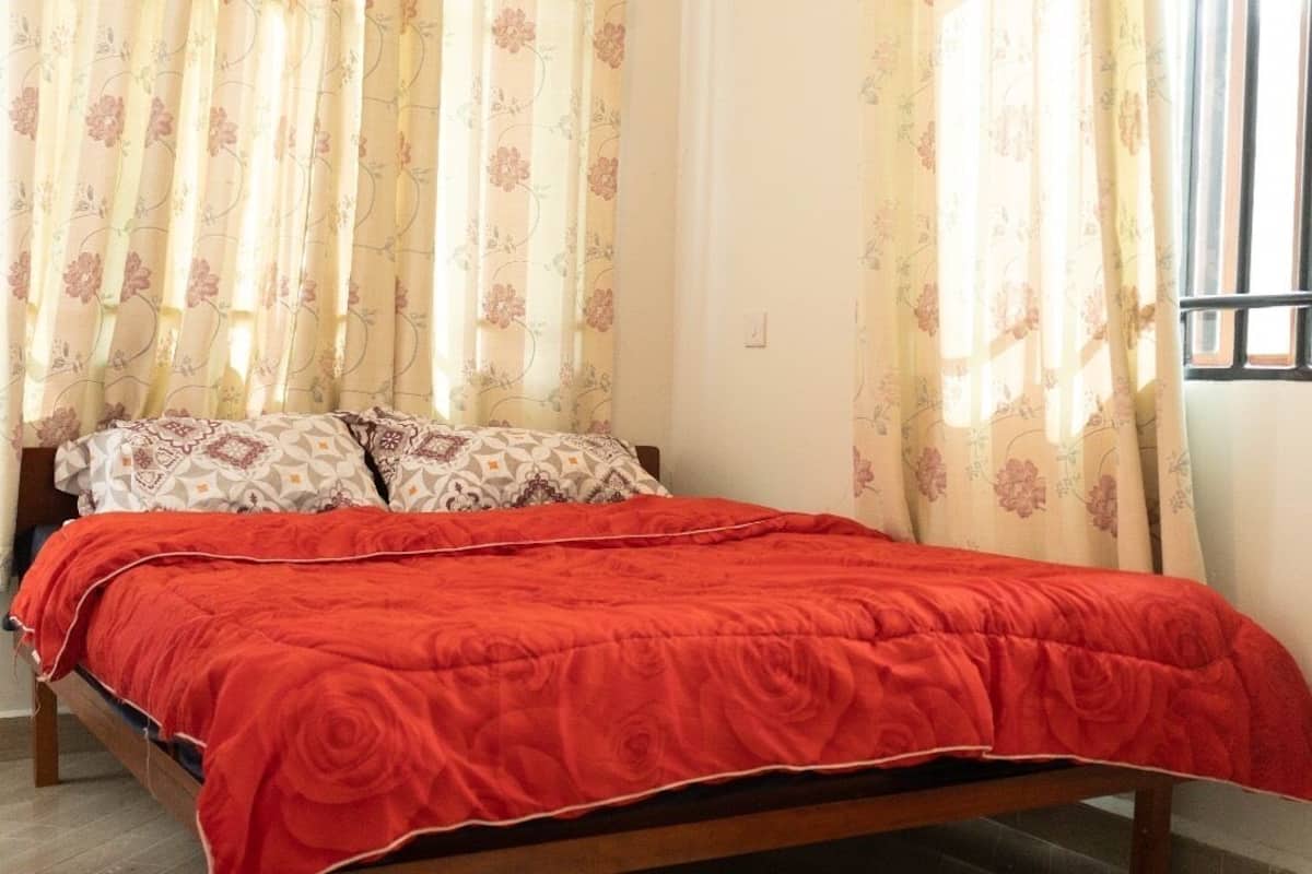 A bed with a red bedspread, floral curtains, and patterned pillows in a room with a window.