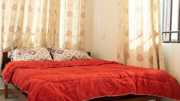 Una cama con una colcha roja, cortinas florales y almohadones estampados en una habitación con ventana.