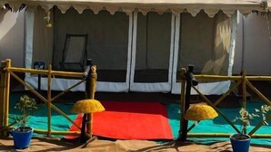 Uma tenda com um colchonete vermelho na frente, cercada por uma cerca de bambu e plantas em vasos no chão.