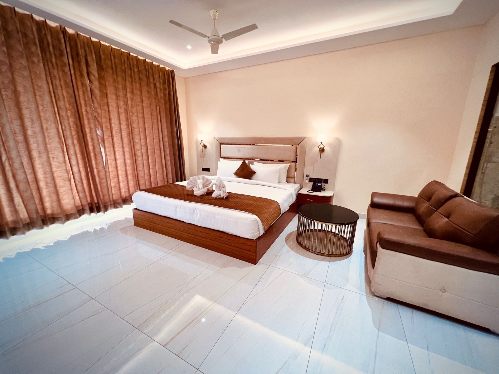 A modern hotel room with a large bed, a round coffee table, a leather sofa, and a ceiling fan.