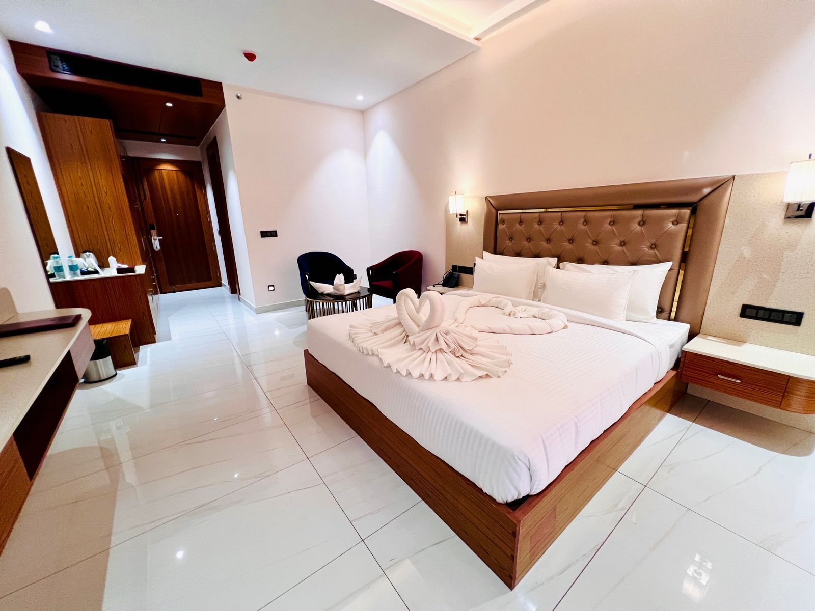 A modern hotel room with a large bed, a wooden headboard, a bedside table, and a desk with a chair.