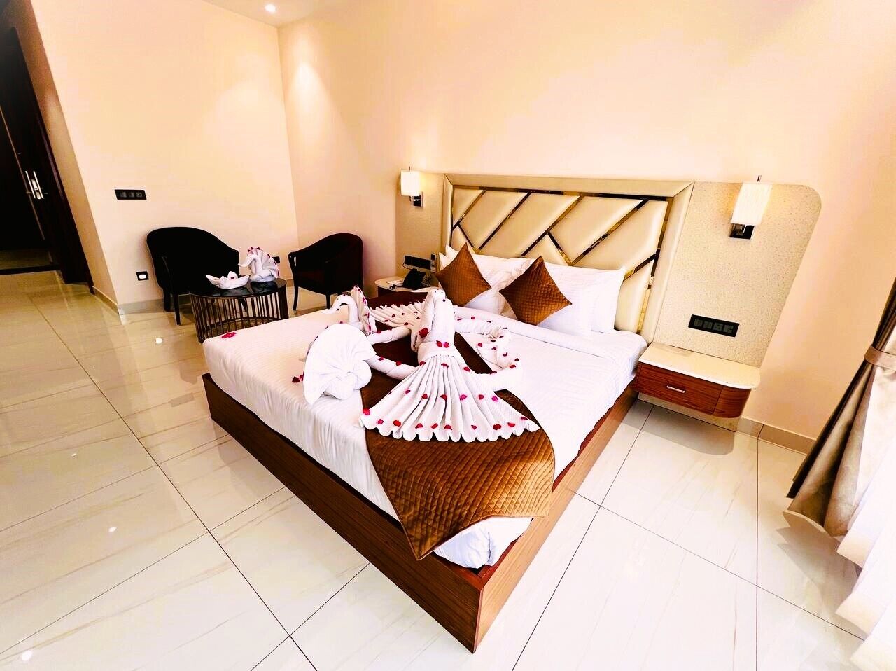 A hotel room with a large bed, a bedside table, a chair, and a small table with a lamp.