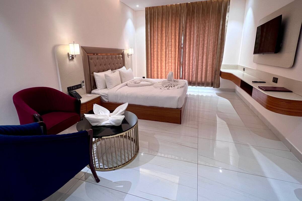 Une chambre d’hôtel moderne avec un grand lit, un fauteuil rouge, une chaise bleue, une table ronde et un téléviseur fixé au mur.