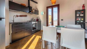 A compact kitchen with a modern stove, oven, and a hanging fish decoration. There is a dining area with a table and chairs, and a large window providing natural light. - Solemar Sicilia - Casa Ernesto  (Santa Flavia)