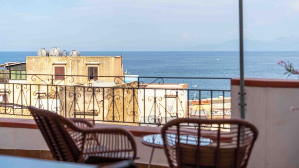 A balcony with wicker chairs, a table, and a view of the sea and buildings. - Solemar Sicilia - Casa Ernesto  (Santa Flavia)