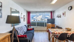 A compact living space with a sofa, a TV, a dining table with chairs, and a view of a snowy landscape through a large window.