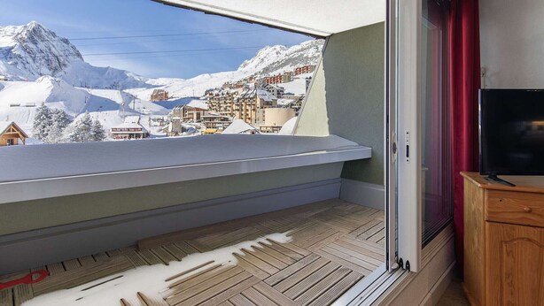A balcony with a view of a snowy mountain town, a wooden floor, and a television on a wooden cabinet.