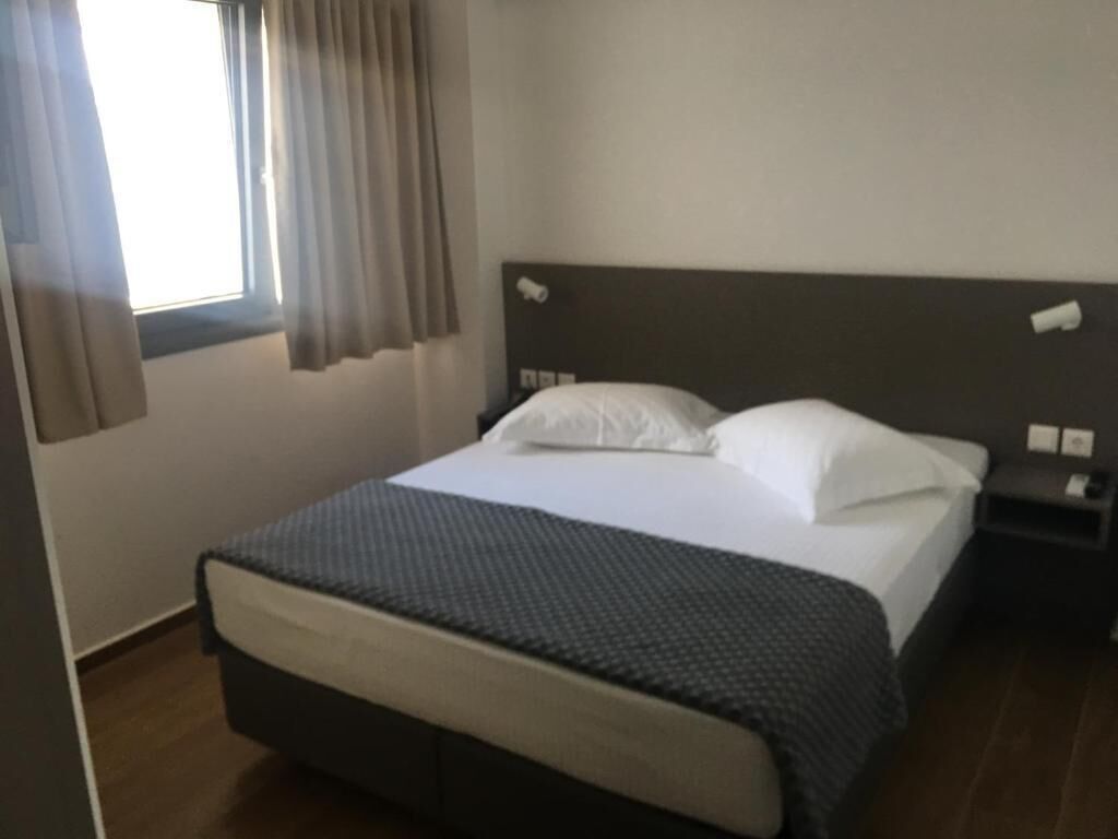 A hotel room with a bed, a nightstand, a window with curtains, and a wall-mounted light switch.