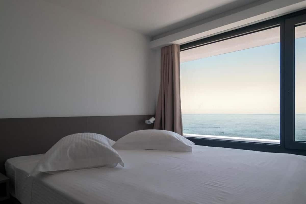 Una habitación de hotel con una cama, una mesita de noche, un ventanal con vistas al mar y una cortina.