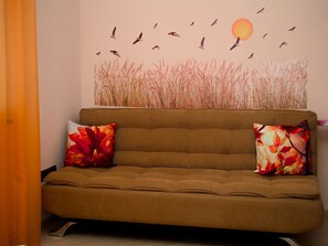A brown sofa with two pillows, a wall with a painting of birds and a sun, and a door with an orange curtain.