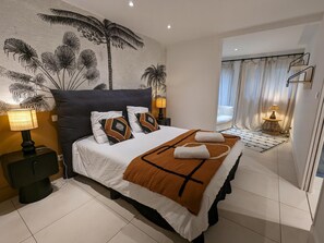 Relax in a spacious room with a large bed, nightstand, and wall-mounted shelf, featuring a palm tree mural.