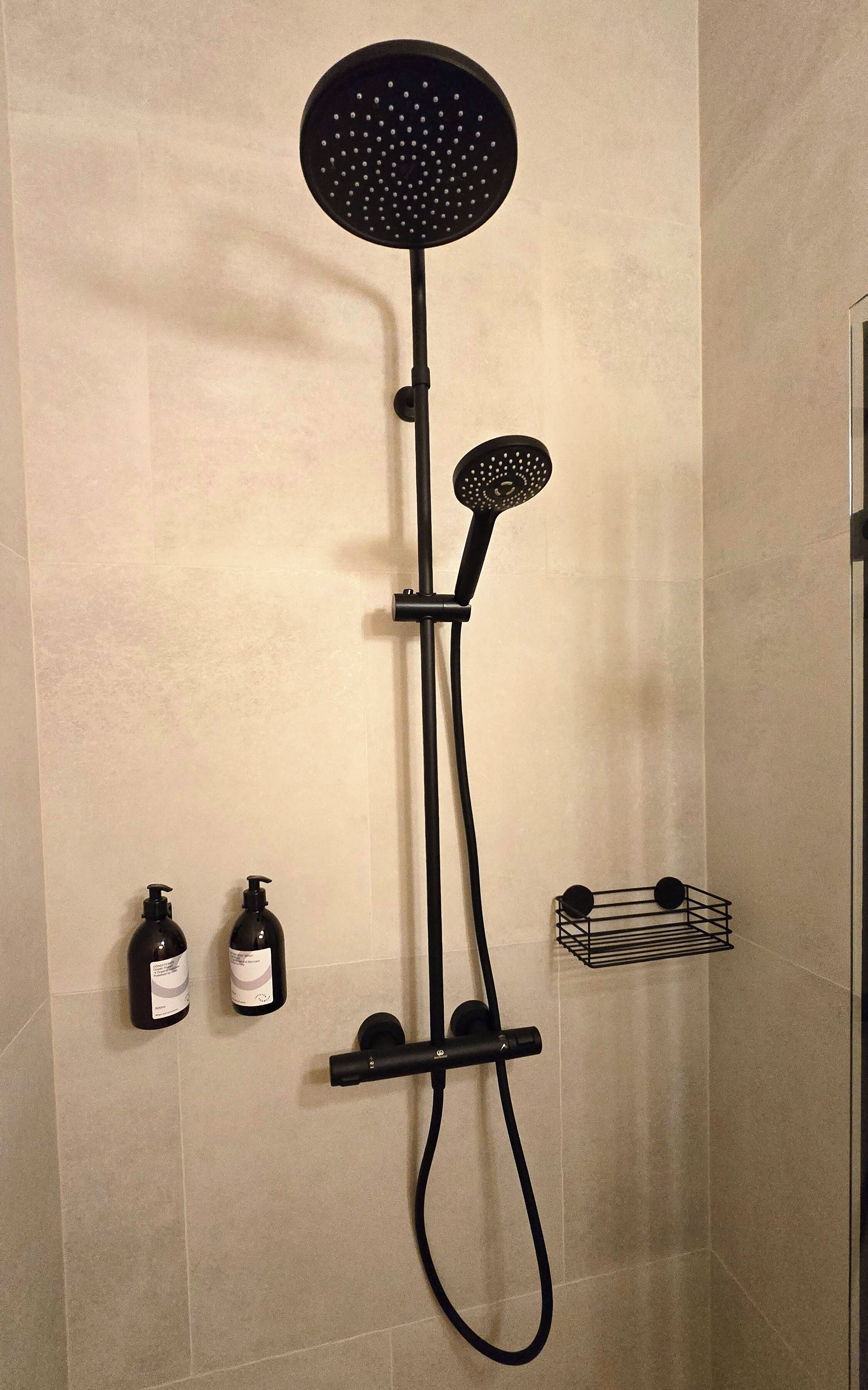 Moderni suihku, jossa on pyöreä suihkupää, käsisuihkupää ja käsisuihkuharja. Seinällä on kaksi pulloa shampoota ja hoitoainetta.