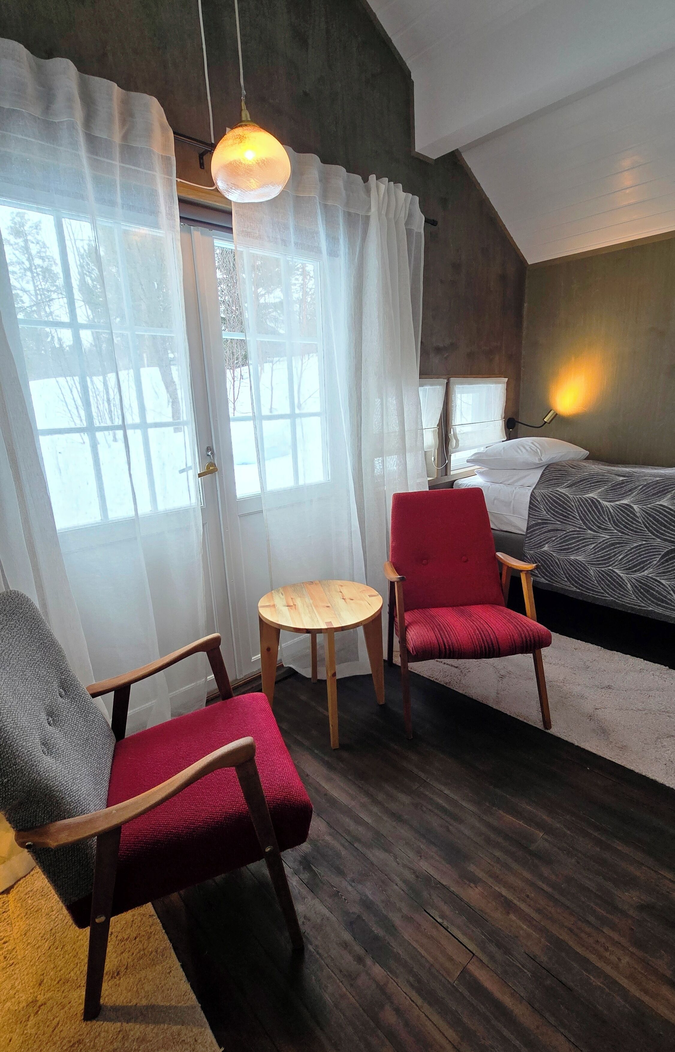 A room with a bed, a red chair, a wooden chair, a small round table, and a hanging light fixture.