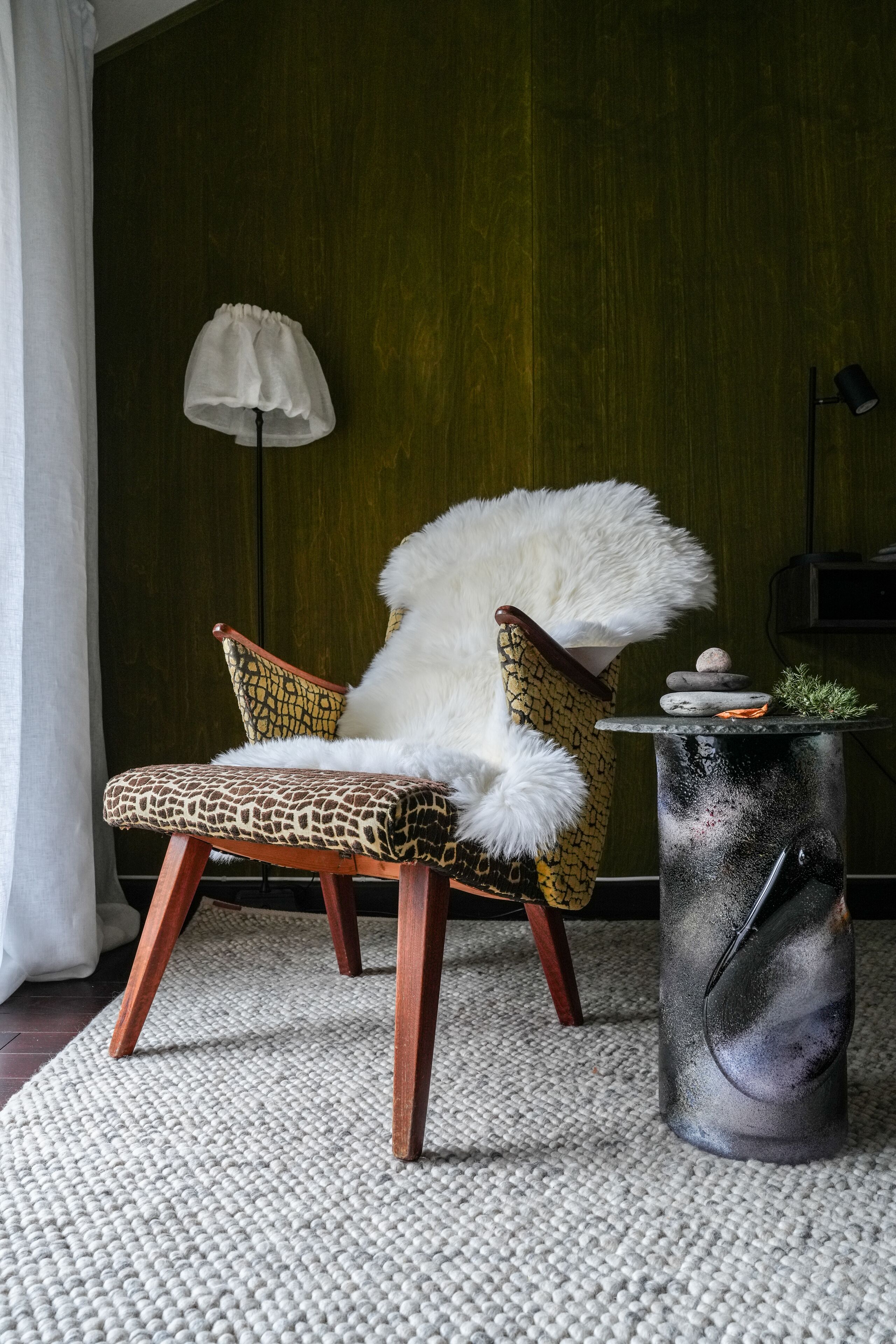 Ein Sessel mit Felldecke, ein kleiner Tisch mit Pflanze und eine Lampe in einem Raum mit grünen Wänden.