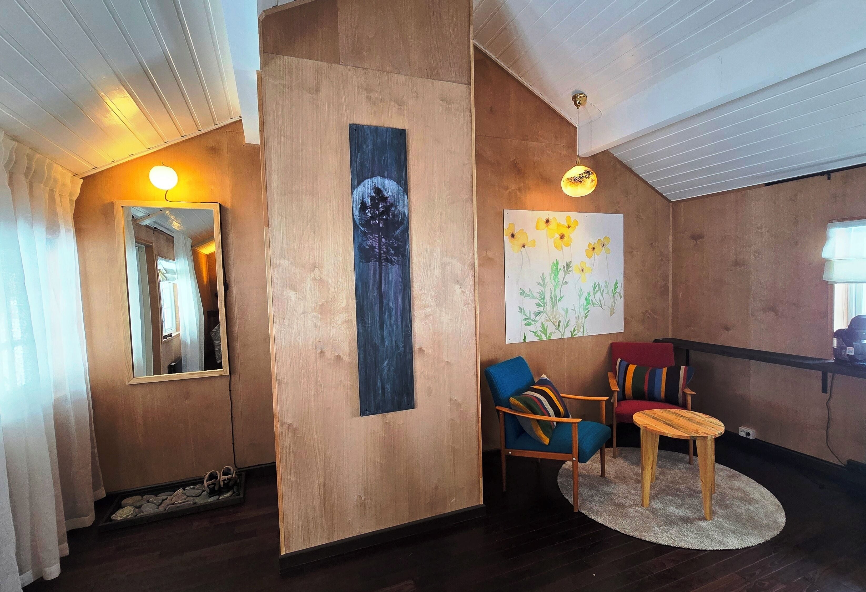 A room with a wooden wall, a mirror, a painting of flowers, two chairs, a small round table, and a light fixture.