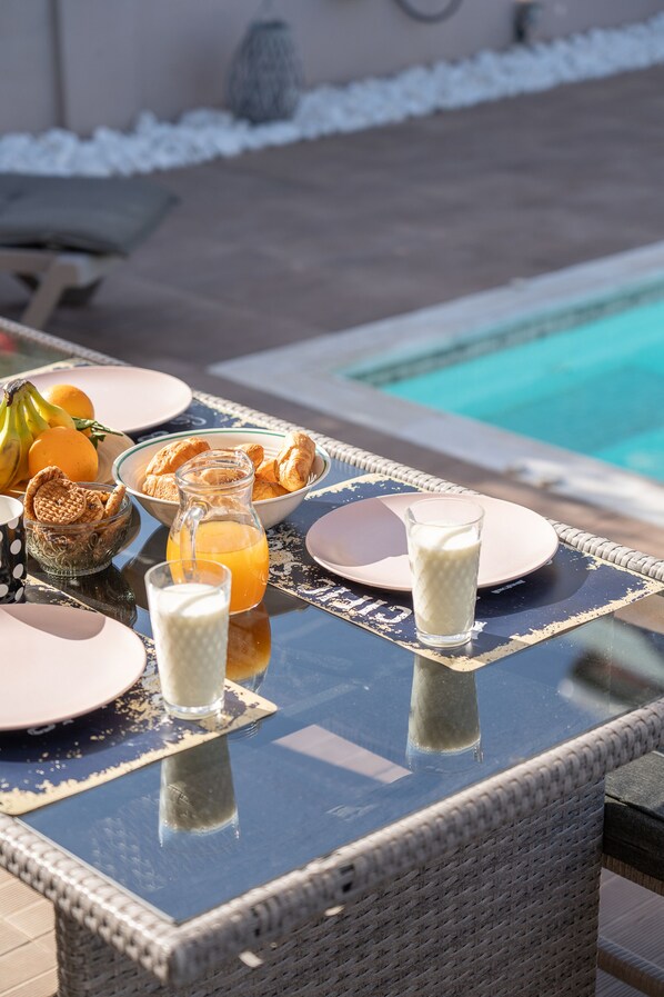 A table with breakfast items including fruits, pastries, and beverages, near a poolside setting.