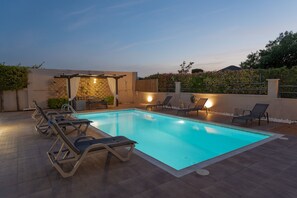 Een rechthoekig zwembad met betegeld zonnedek, omringd door ligbedden en een pergola met verlichting.