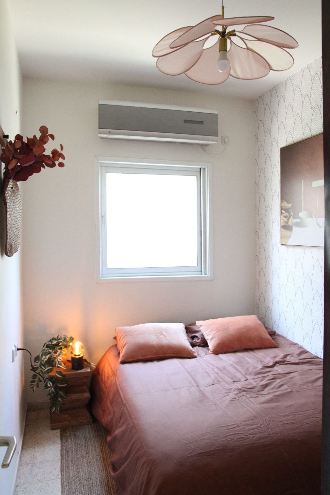 A bedroom with a bed, a window, an air conditioning unit, a bedside lamp, and a wall-mounted picture.