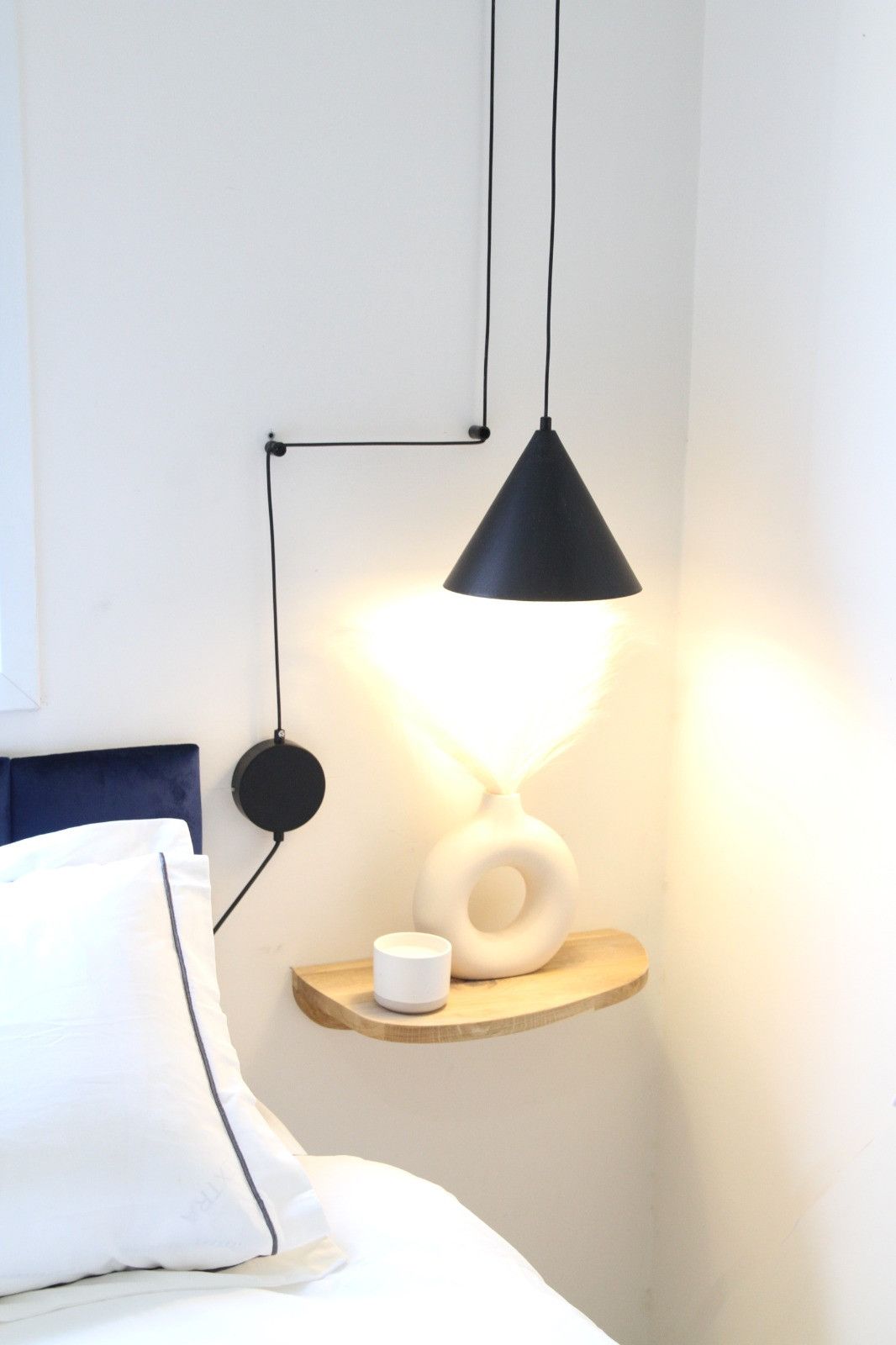 A bed with white bedding, a white pillow, and a bedside table with a white vase and a white cup.