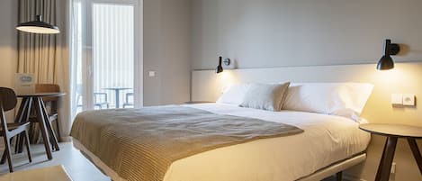 A hotel room with a large bed, a bedside table, a chair, and a small table with a lamp.