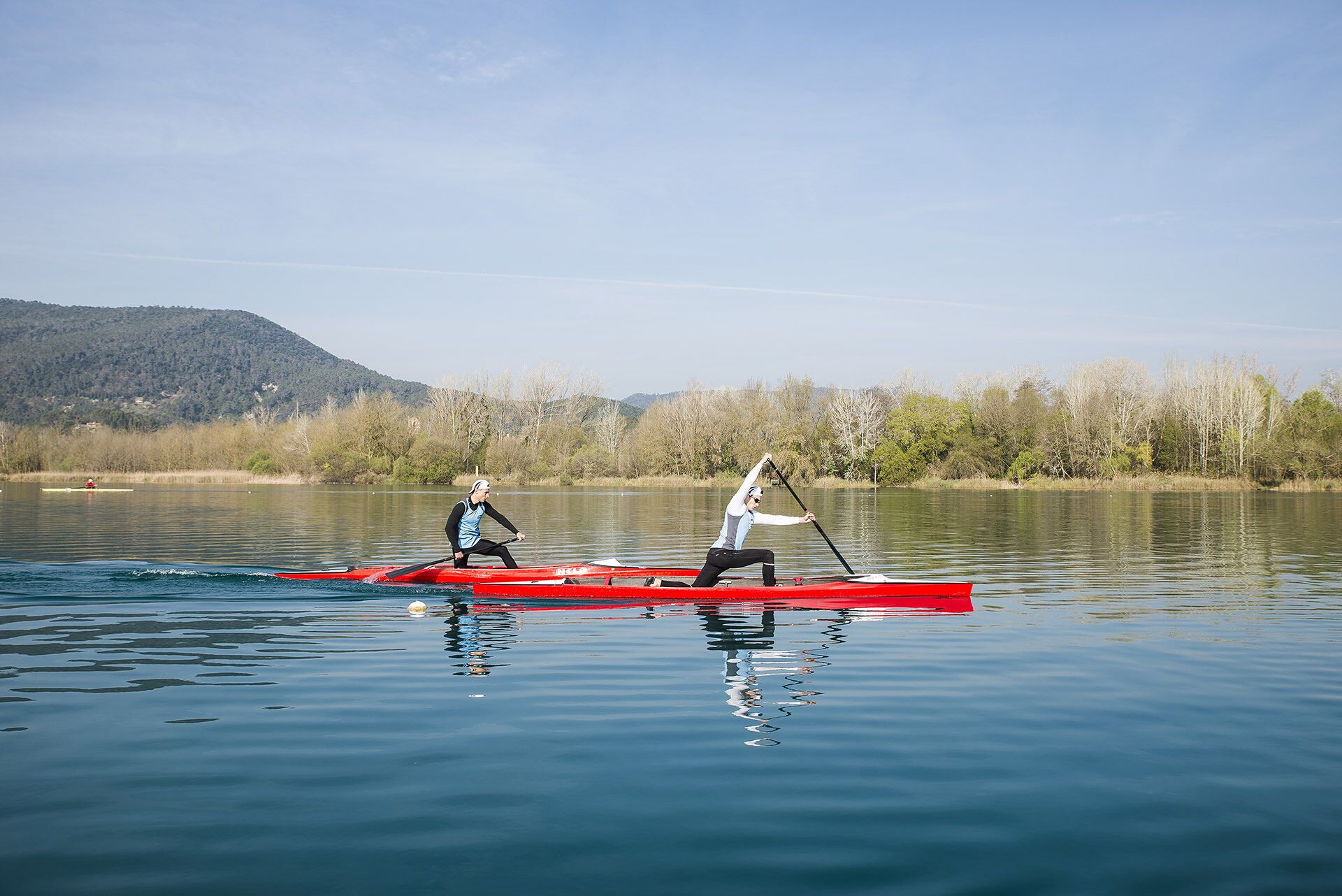Dues persones remen a una caiac vermella sobre un llac tranquil, amb un vessant boscos en el fons.